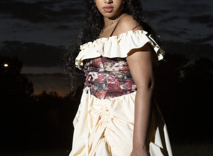 Model @mocha_bunn wearing a cream off-the-shoulder dress with a burgundy floral corset, posing beneath a dark twilight sky at Ray Trent Park in Midwest City, Oklahoma.