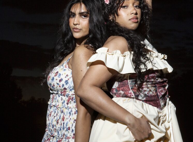 Models @alaya.jaffery in a flowing floral sundress and @mocha_bunn in a cream off-the-shoulder dress with a burgundy floral corset, posing back-to-back with raised arms against a dark evening sky at Ray Trent Park.