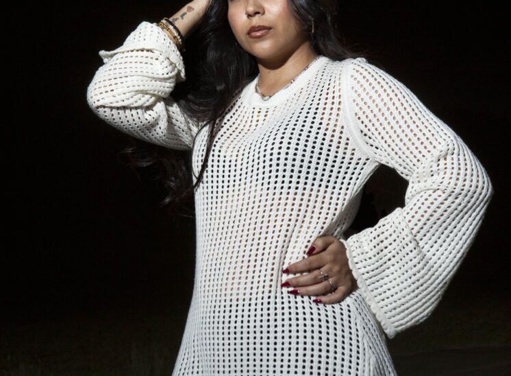 Model @_cilllaaaa wearing a white open-knit dress, posing confidently with one hand resting on her head and the other on her hip against a dark nighttime background at Ray Trent Park.